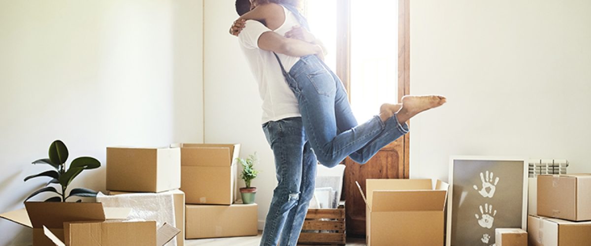 Full length of young man lifting woman in new apartment. Loving couple is surrounded with cardboard boxes. They are wearing casuals.